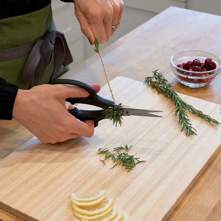 Hands using Shun Herb Shears to strip rosemary leaves from the stem on a cutting board.
