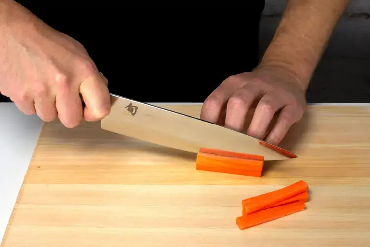 Chef using a Shun Classic Chef's knife to slice carrots into batonnet sticks on a hinoki cutting board.