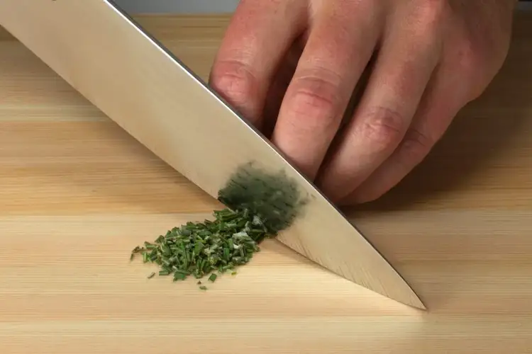 Close-up of a Shun knife mincing fresh herbs on a wooden cutting board with a fine rocking motion.