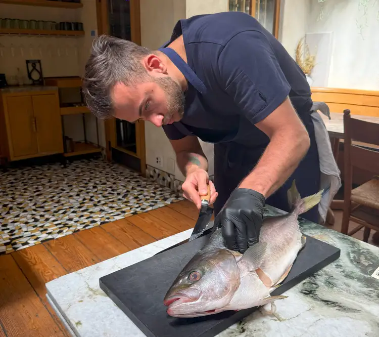 Chef Vitor Mendes filleting a whole fish with precision using a Shun Boning & Fillet knife on a marble counter at Theodora in Brooklyn. He is wearing a navy apron and black gloves, focused intently on his technique.