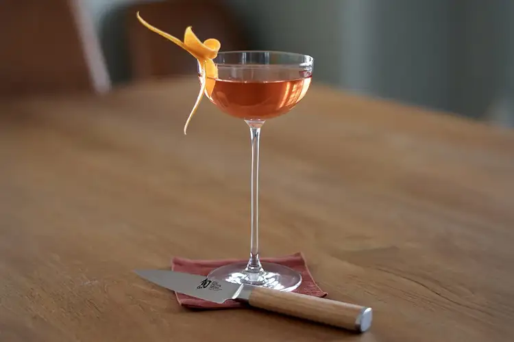 A finished coctail with beautiful orange peel garnish. The Classic Blonde Paring Knife sits in front of the glass.