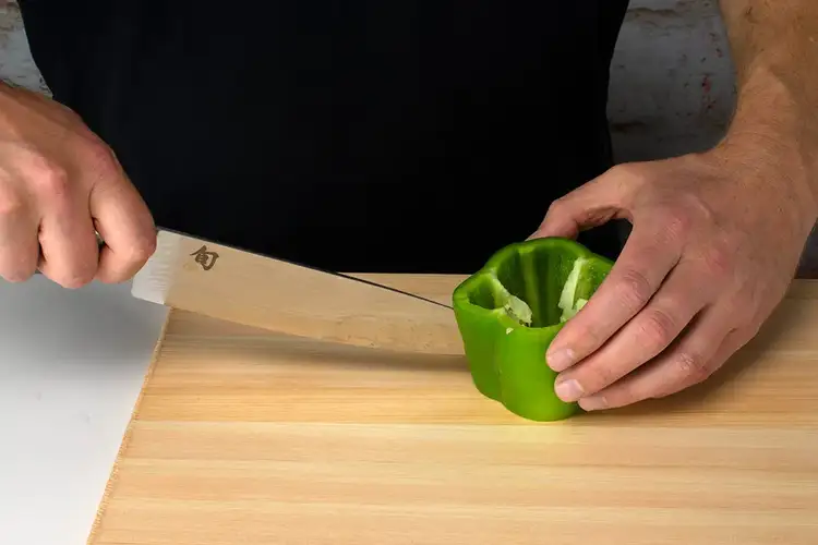 The top of the bell pepper is removed, exposing the inner seeds; the knife is poised to slice down the side.