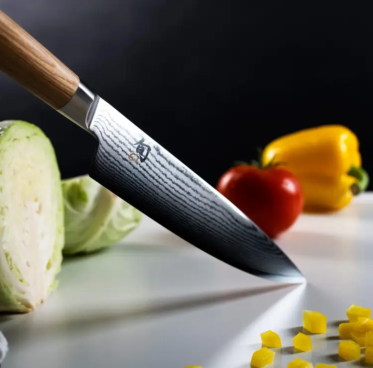 Shun Classic Blonde Chef’s Knife with Damascus-patterned blade resting beside fresh vegetables, highlighting precision design and light-colored Pakkawood handle. Click to shop this knife.