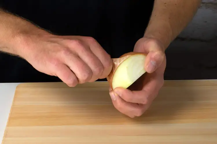 Hands peeling away the outer skin of the onion after slicing in half to prepare it for chopping.