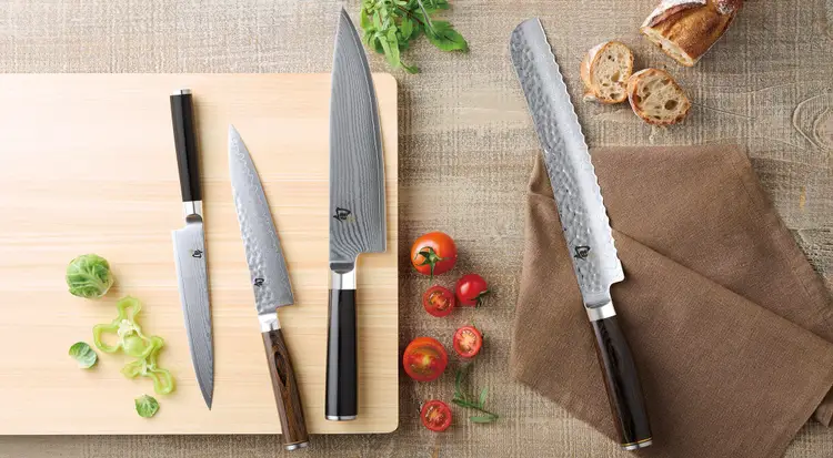 Shun Japanese knives arranged on a Hinoki cutting board with fresh vegetables and bread, demonstrating proper cutting surfaces and knife care.
