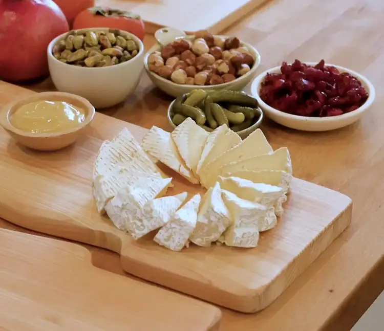 Camembert wedges fanned into a flower shape on a board, surrounded by bowls of nuts, mustard, and pickles.