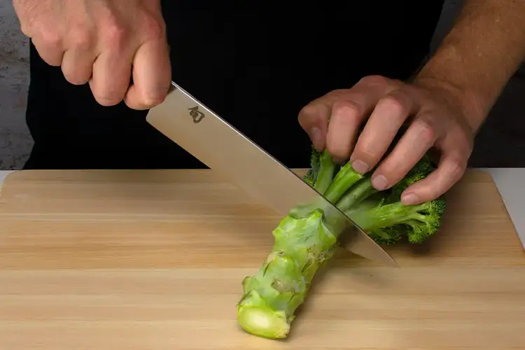 The hand holds the broccoli firmly as the knife slices further through the stalk, separating florets.