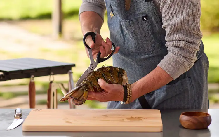 Hands using Shun kitchen shears to cut through a lobster tail on a cutting board.
