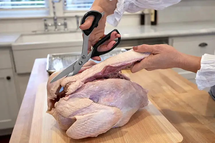 Hands using Shun kitchen shears to remove the backbone from a whole chicken on a cutting board.