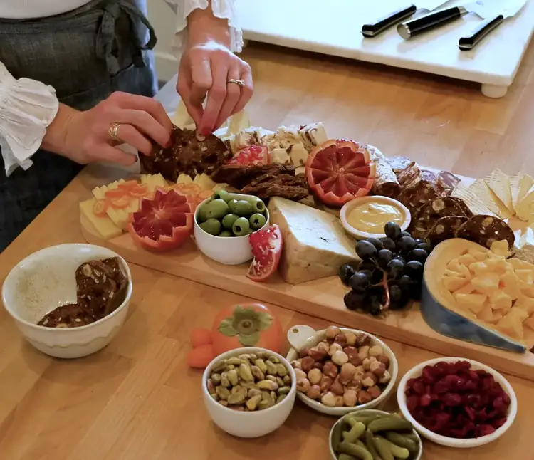 Placing crackers on a colorful cheeseboard filled with sliced cheese, olives, and garnished fruits like blood orange and pomegranate.