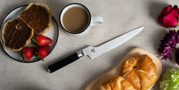 Shun Classic Offset Bread Knife rests on a table surrounded by breakfast pastries and flowers. Click to shop the Classic Offset Bread Knife.