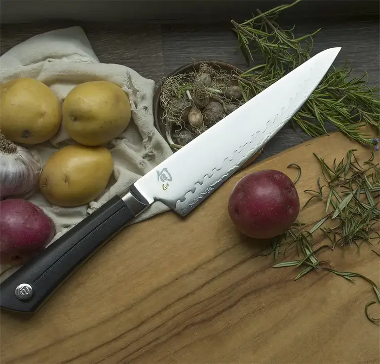 Shun Sora Chef’s Knife placed on a wooden board with potatoes, garlic, and rosemary, styled to evoke a rustic meal prep setting.