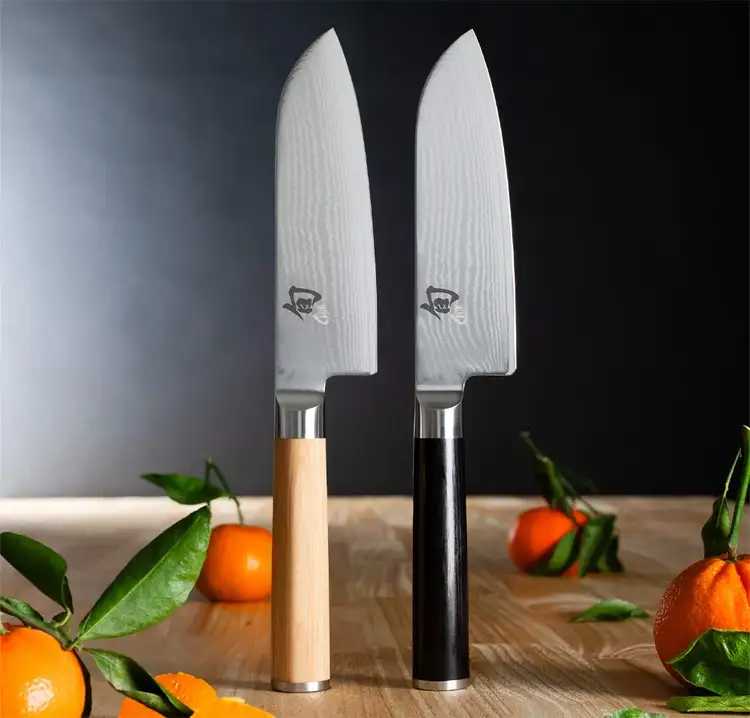 Image of two Classic Santokus, one with an ebony and one with a blonde handle, standing vertically on a table.