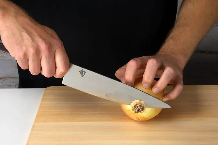 Chef trimming the top of a halved onion with a Shun chef’s knife while keeping the root end intact for stability.
