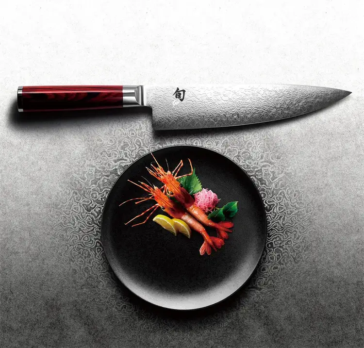 A dramatically lit Kohen Chef's Knife rests above a plated shrimp dish.