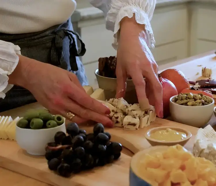 Arranging cranberry-style flavored cheese chunks on a cheeseboard surrounded by olives, grapes, and other snacks.