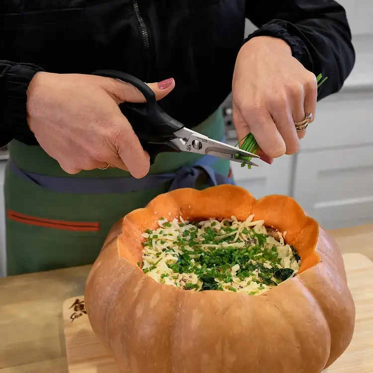 Hands using kitchen shears to cut fresh chives over a stuffed squash on a cutting board.