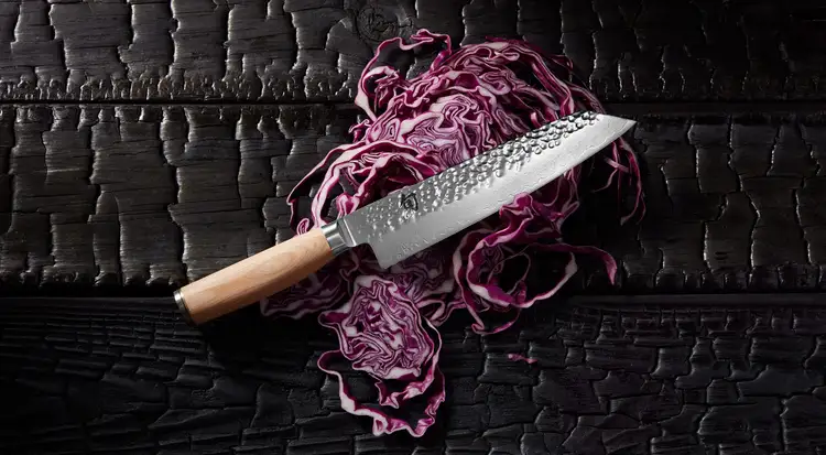 Shun Premier Blonde Kiritsuke knife resting on thinly sliced red cabbage atop a charred wood surface. Click to shop.