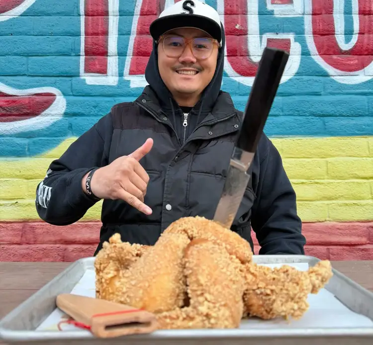 Chef Paolo Campbell smiles in front of a colorful mural, holding a tray of crispy fried chicken with a Shun knife dramatically placed in the center.