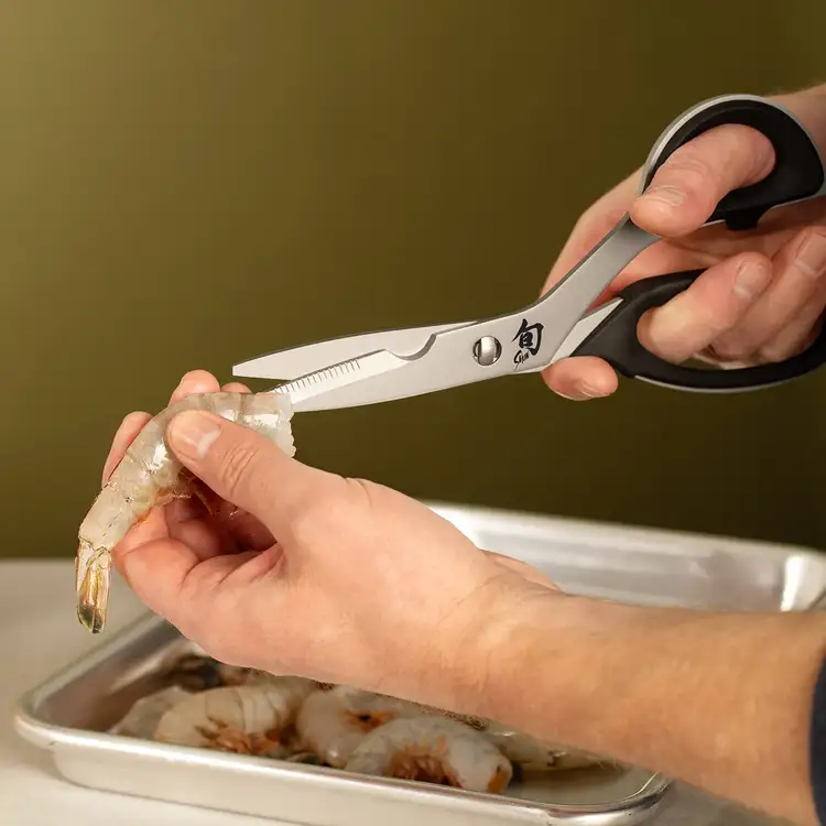 Person using Shun kitchen shears to trim raw shrimp over a metal tray