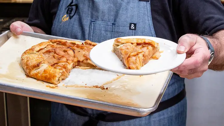 Andrew holding an Apple Galette on a cooking sheet with his left hand. In his other hand Andrew holds a slice of the Apple Galette on a small white plate. Click to play video