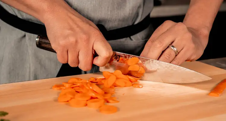 Close-up of a chef slicing carrots with a Shun Premier Chef’s Knife, showcasing its hammered tsuchime finish and razor-sharp edge on a wooden cutting board. Click to shop this knife.