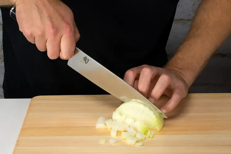 Chef finishing a fine dice of onion with smooth, downward cuts, showing uniform pieces on the board.