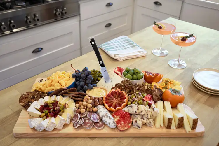 A fully styled cheese board with a variety of cheeses, fruits, nuts, and salami, with a Shun Kazahana utility knife standing in a wedge of blue cheese.