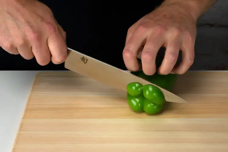 A hand begins slicing the top off a green bell pepper using a Shun Classic Chef's Knife on a wood cutting board.