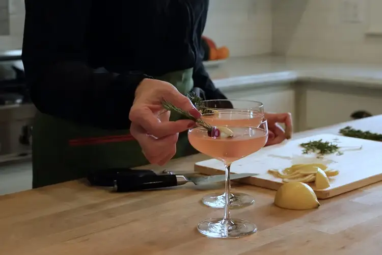 A festive rosemary and cranberry garnish being placed into a pink holiday cocktail.