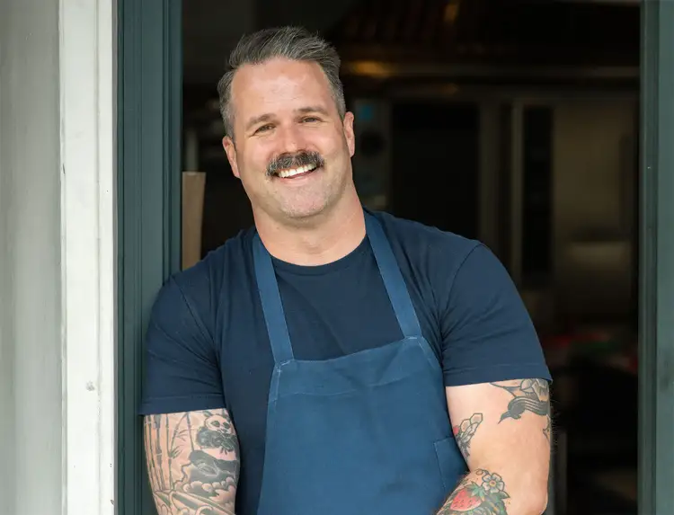 Chef Avery Hardin standing in a kitchen doorway, wearing an apron and smiling.