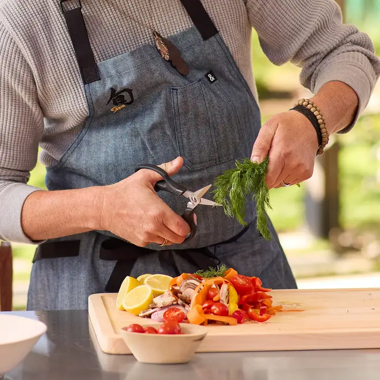 Kitchen shears cutting fresh herbs over a cutting board with prepared vegetables nearby.