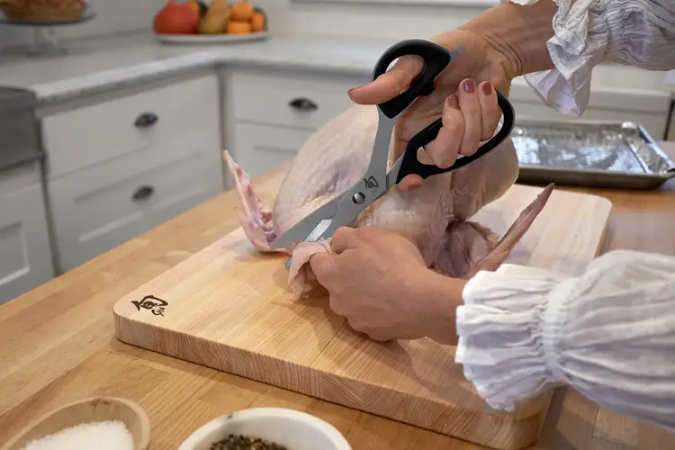 Trimming excess skin from a raw turkey using Shun Kitchen Shears on a wooden Hinoki cutting board.