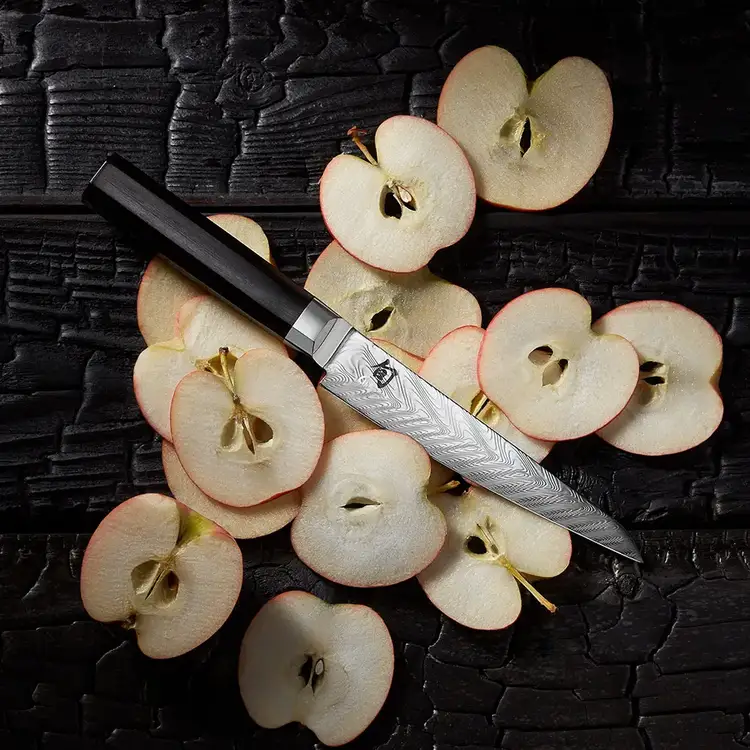 A Dual Core Utility/Butcher knife lices on thin slices of apple spread across a table. Click to shop.