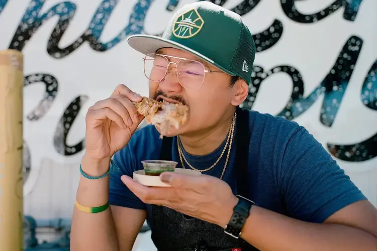 Chef Paolo Campbell enjoying a crispy fried chicken drumstick, dipping it into a small bowl of sauce outside The Chicken Supply.