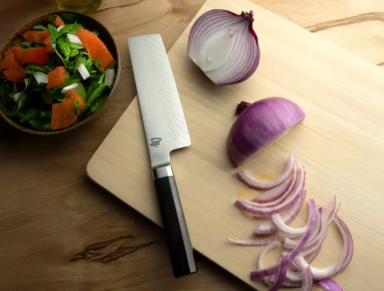 Shun Dual Core Nakiri knife on a hinoki cutting board beside a halved red onion. Click to shop this knife.