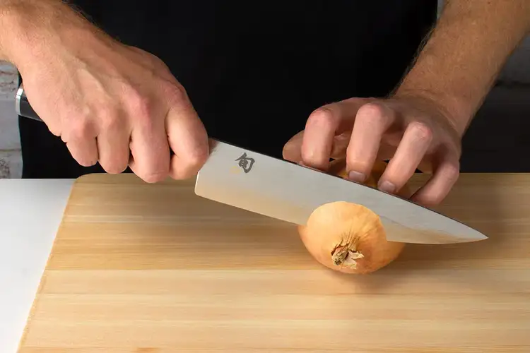Chef slicing an onion in half vertically through the root end using a Shun chef’s knife on a hinoki cutting board.