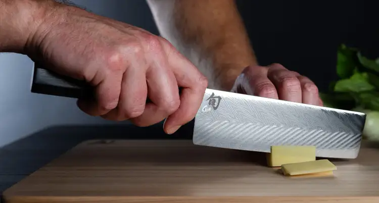 Chef slicing ginger with a Shun Dual Core Nakiri knife on a hinoki cutting board, showcasing the layered Damascus pattern and flat blade profile.