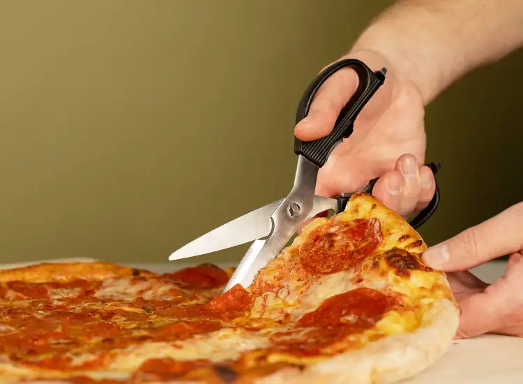 Kitchen shears cutting a slice of pepperoni pizza on a cutting board.