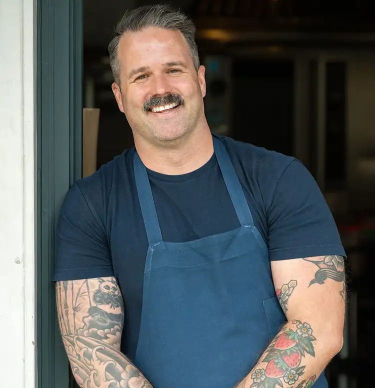 Chef Avery Hardin standing in a kitchen doorway, wearing an apron and smiling. Click to read his interview.