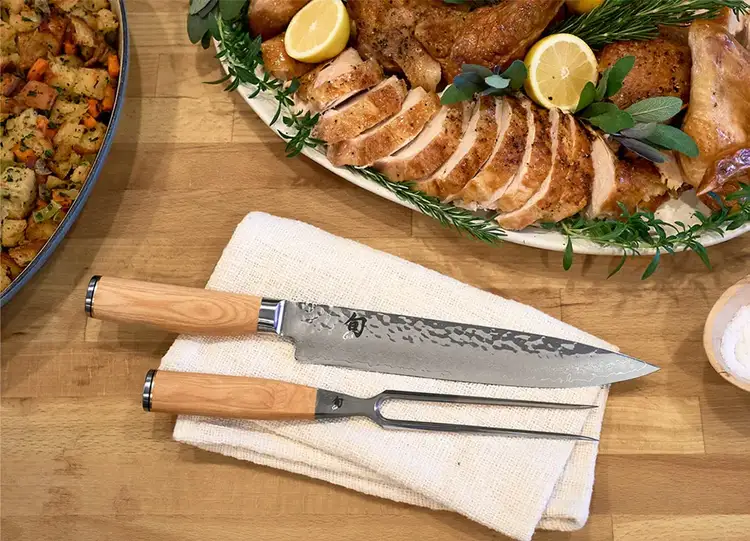 A Premier Blonde slicing knife and carving fork stand on a table next to a holiday feast.