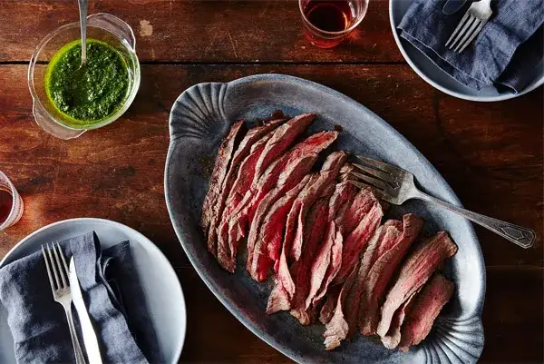 Sliced flank steak on platter with green sauce in a bowl