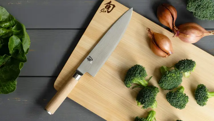 Shun Classic Blonde Chef’s knife resting on a hinoki cutting board with broccoli florets and shallots. Click to shop this knife.