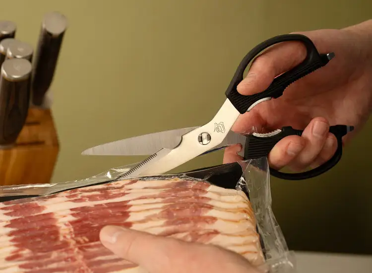Kitchen shears opening plastic packaging on sliced bacon.