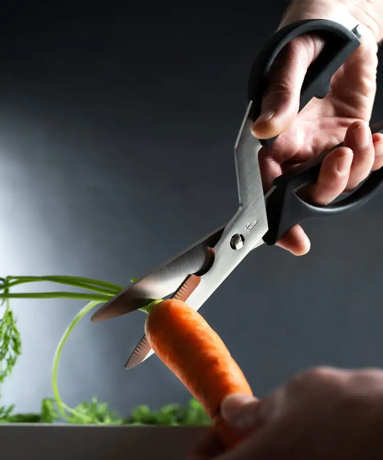 Close-up of kitchen shears trimming a carrot stem over a bowl.